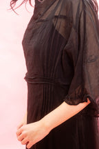 A close-up of a person wearing a black sheer mesh overlay dress with a gathered waist and ruffled elbow-length sleeves, set against a solid pink background.