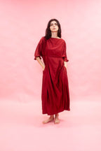 A woman stands barefoot against a solid pink studio background, wearing a loose, long-sleeved maxi dress in deep burgundy red with side pockets.