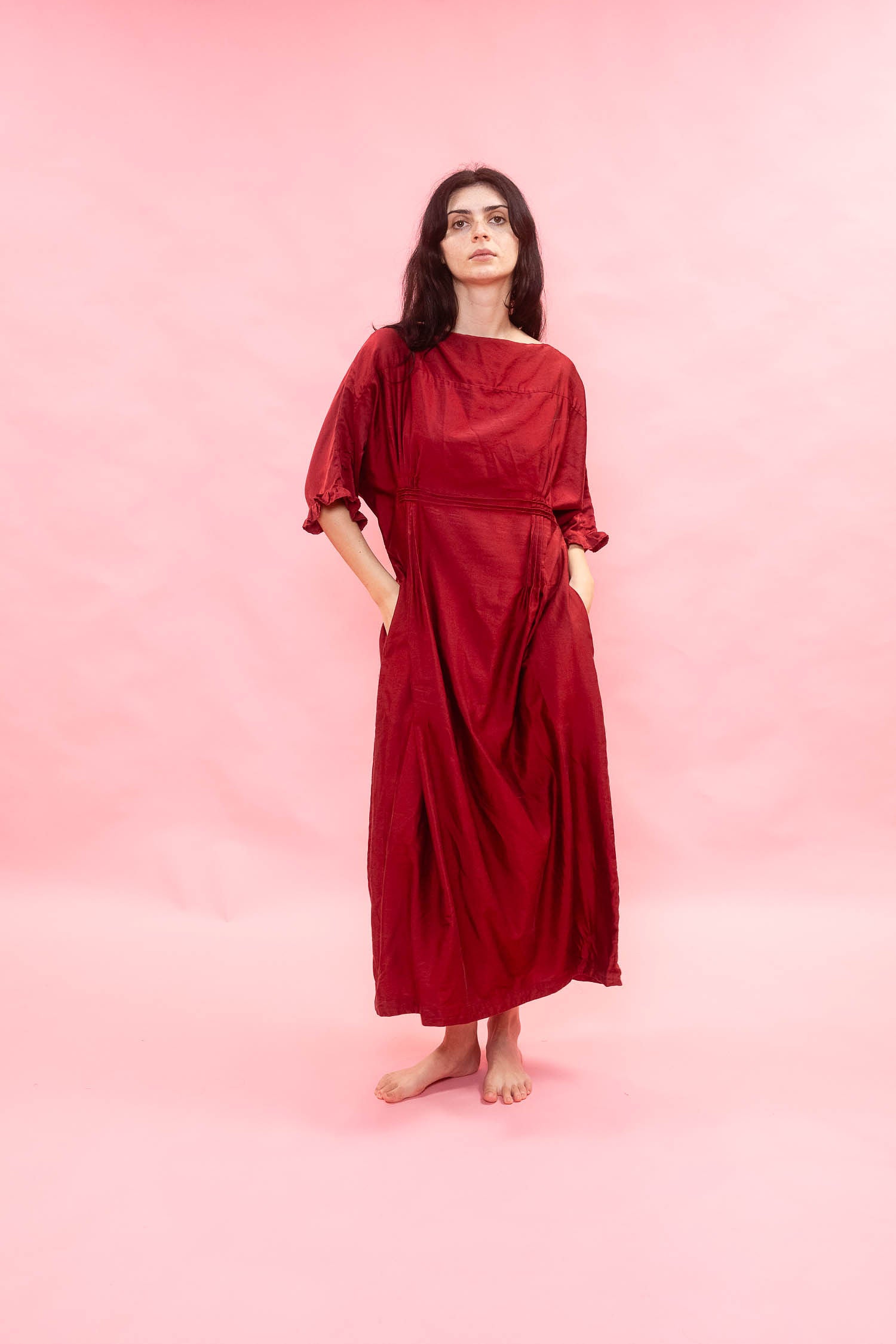 A woman stands barefoot against a solid pink studio background, wearing a loose, long-sleeved maxi dress in deep burgundy red with side pockets.