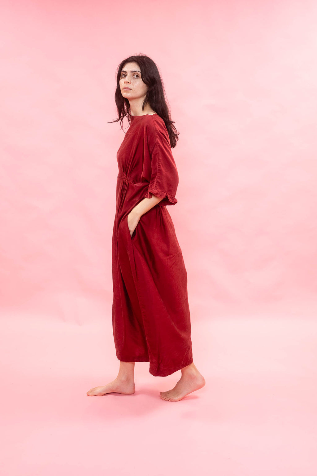 A woman stands barefoot in profile wearing a loose, long-sleeved red dress against a solid pink studio background.