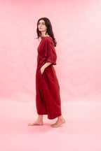 A woman stands barefoot in profile wearing a loose, long-sleeved red dress against a solid pink studio background.