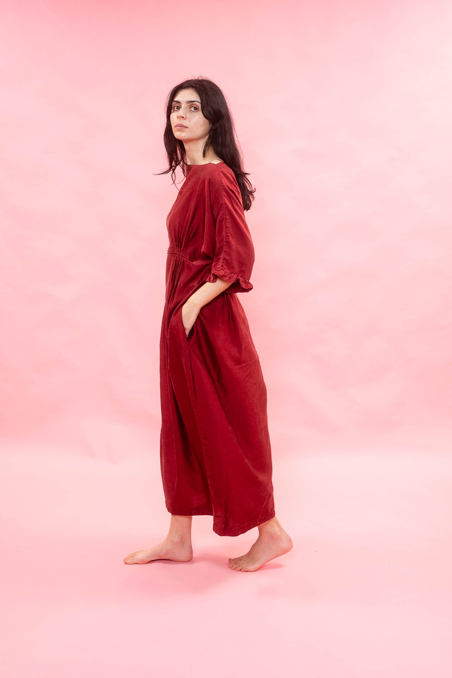 A woman stands barefoot in profile wearing a loose, long-sleeved red dress against a solid pink studio background.