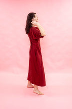 A woman wearing a long, dark red dress stands barefoot against a solid pink studio background, posing in profile.