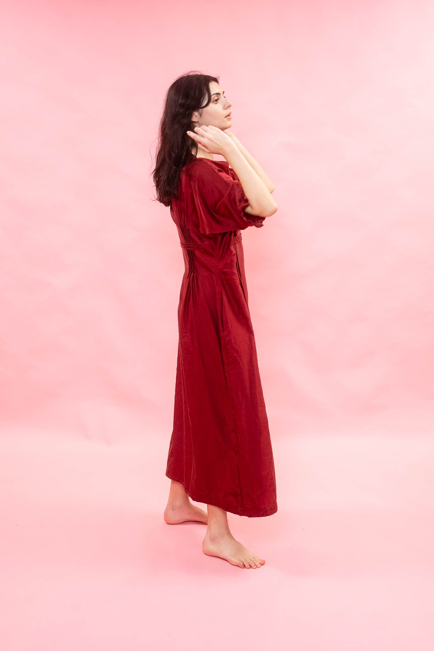 A woman wearing a long, dark red dress stands barefoot against a solid pink studio background, posing in profile.