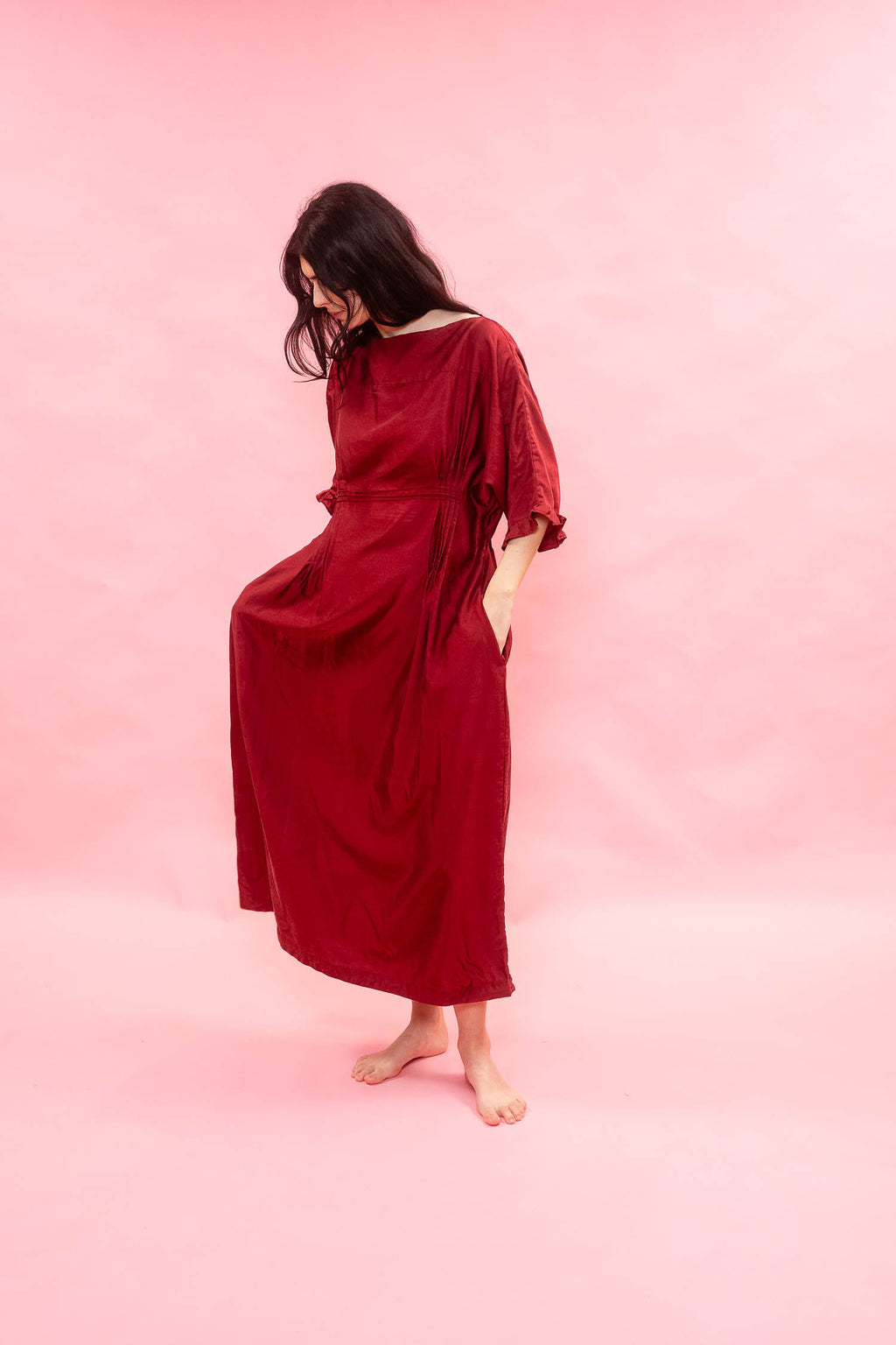 A woman wearing a loose, long-sleeved red maxi dress with side seam pockets, standing barefoot against a solid pink backdrop.