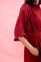 A close-up side view of a woman wearing a loose, dark red dress with elbow-length sleeves with a frill hem and a pink tucked waist, posed against a solid pink background.