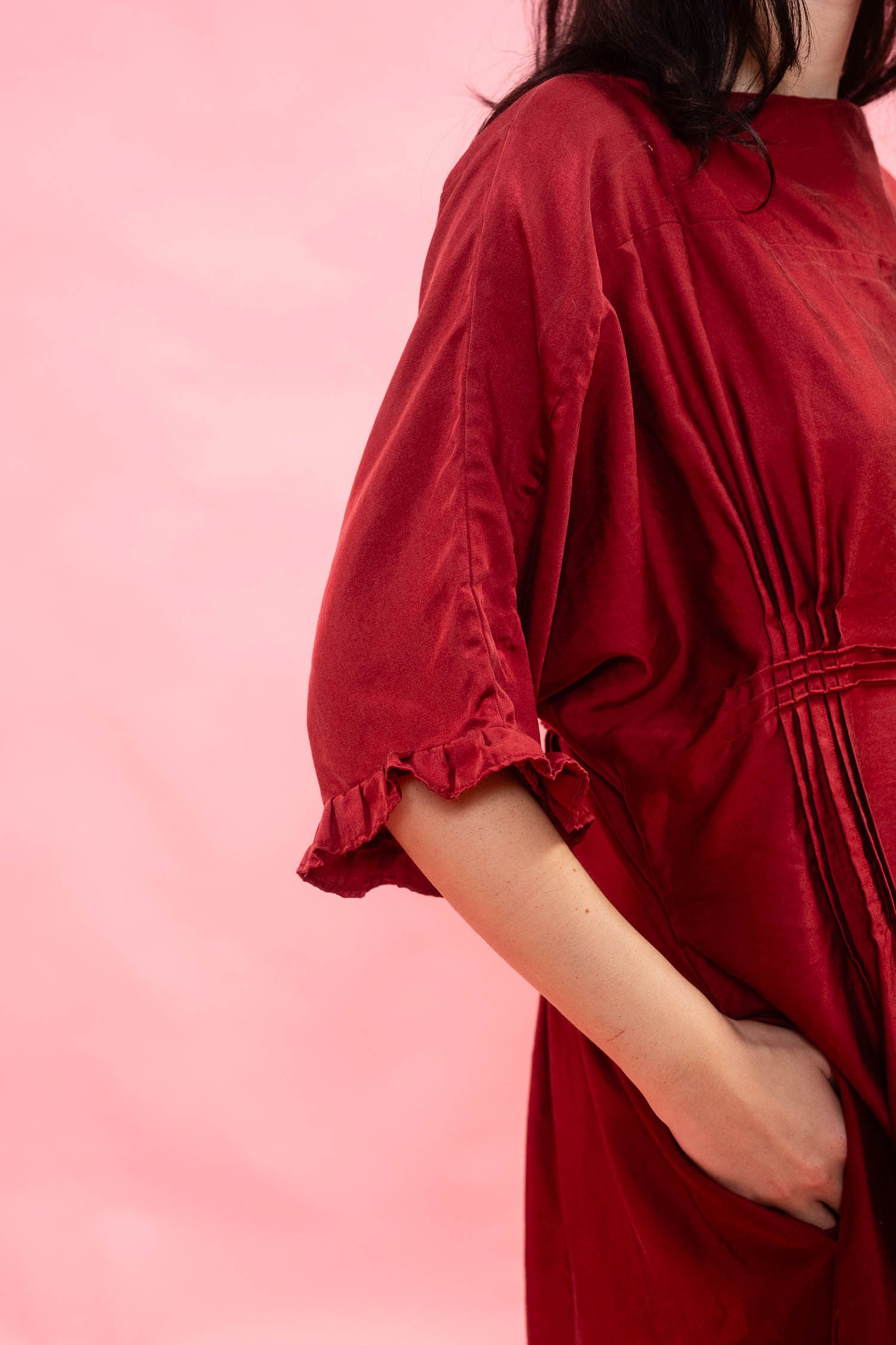 A close-up side view of a woman wearing a loose, dark red dress with elbow-length sleeves with a frill hem and a pink tucked waist, posed against a solid pink background.