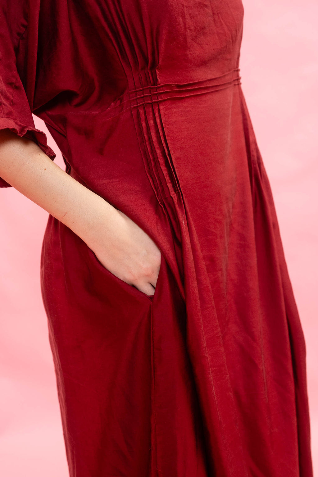 A close-up of a woman wearing a deep red maxi dress with pleated details and a side pocket, with their hand in the pocket, set against a solid pink background.