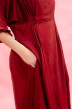 A close-up of a woman wearing a deep red maxi dress with pleated details and a side pocket, with their hand in the pocket, set against a solid pink background.