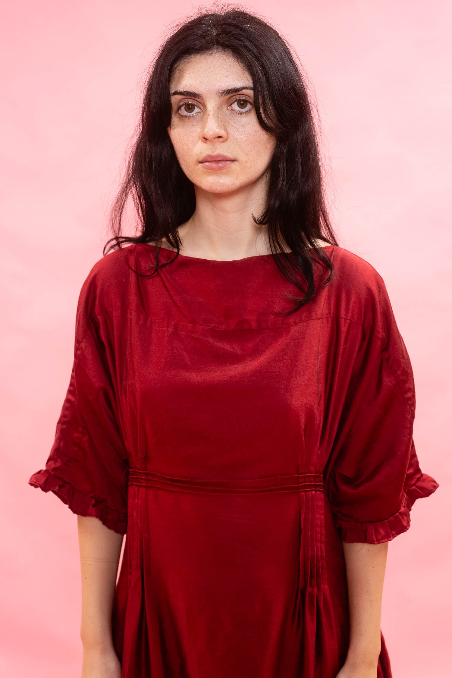 A woman with dark hair wearing a deep red dress with three-quarter wide relaxed sleeves, standing against a solid pink background.