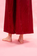 A close-up of a woman's bare feet and lower legs wearing a red silk satin midi length dress against a solid pink background.