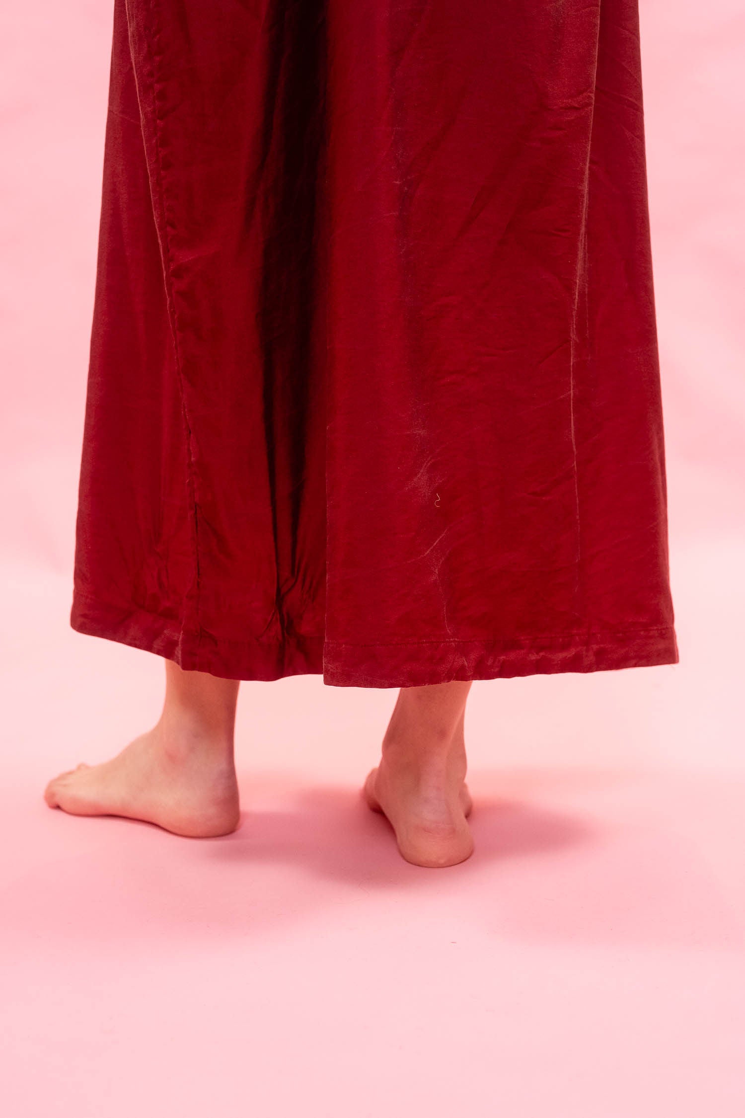 A close-up of a woman's bare feet and lower legs wearing a red silk satin midi length dress against a solid pink background.