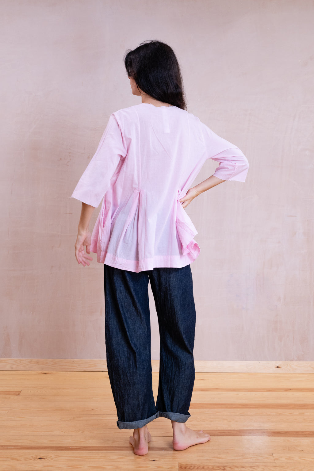 A woman viewed from behind wearing a light pink short-sleeve peplum-style top and dark blue denim pants, standing on a light wood floor against a textured beige wall.