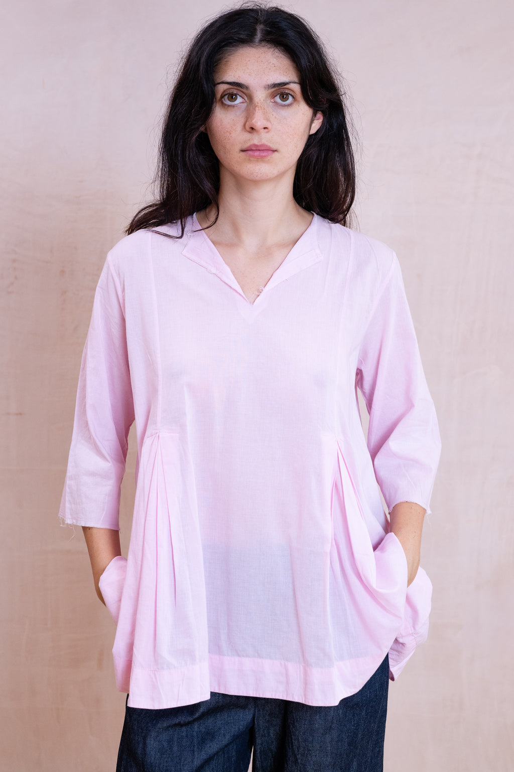 A woman wearing a light pink V-neck tunic top with three-quarter sleeves and side pockets, paired with dark blue jeans, standing against a textured beige background.