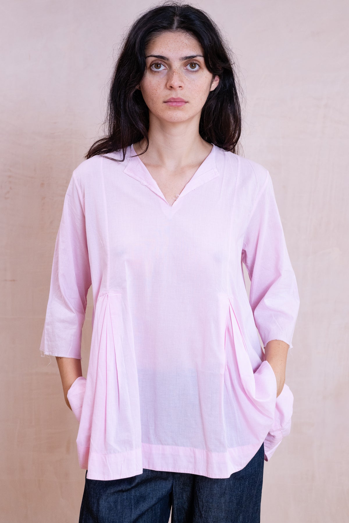 A woman wearing a light pink V-neck tunic top with three-quarter sleeves and side pockets, paired with dark blue jeans, standing against a textured beige background.