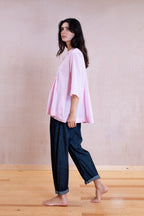 A woman wearing a light pink oversized short-sleeve top and dark blue cropped jeans, standing barefoot on a light wood floor against a textured beige wall.
