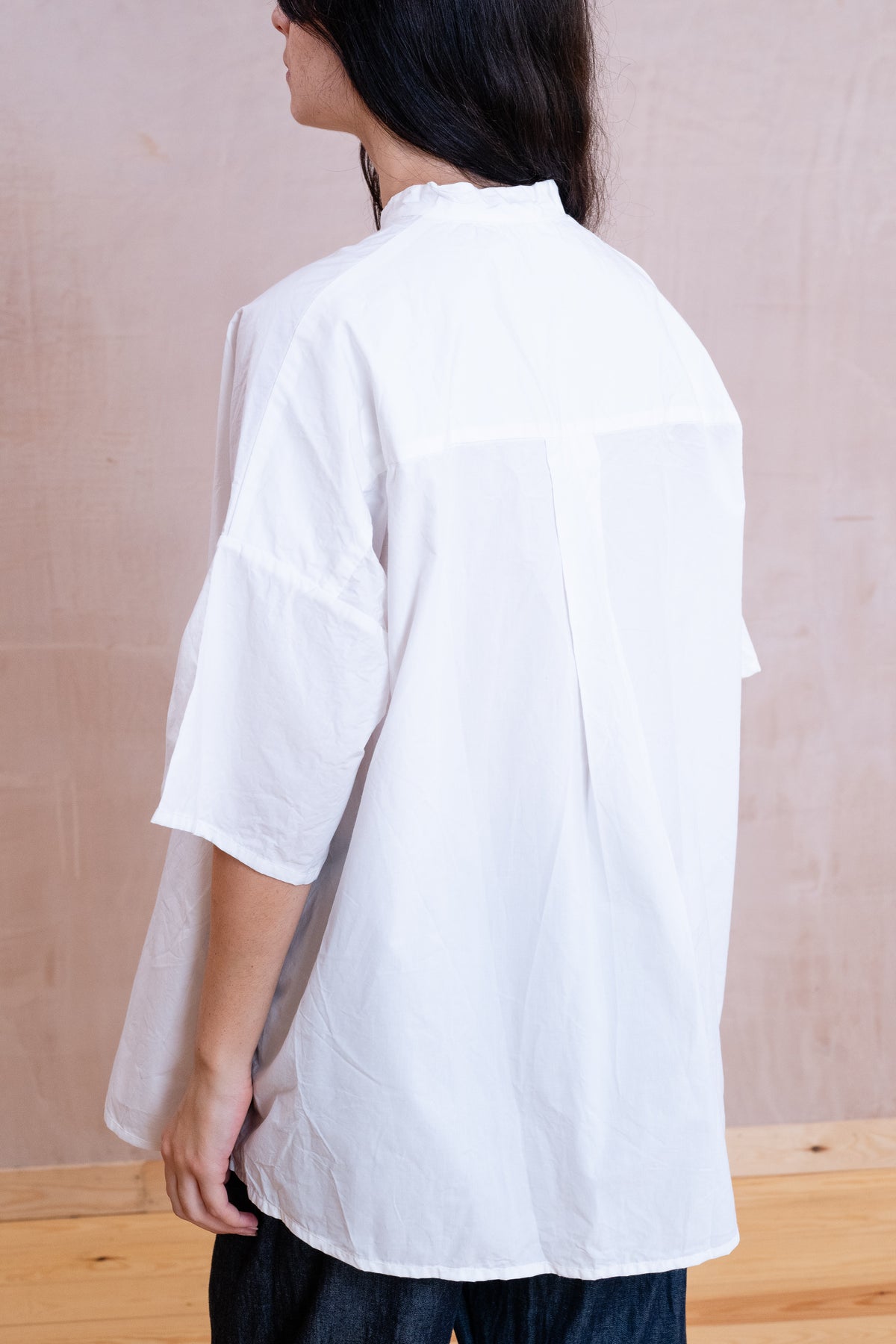 A person shown from the back wearing an oversized white short-sleeve shirt with a high collar and dark pants, standing against a light beige wall and wooden floor.