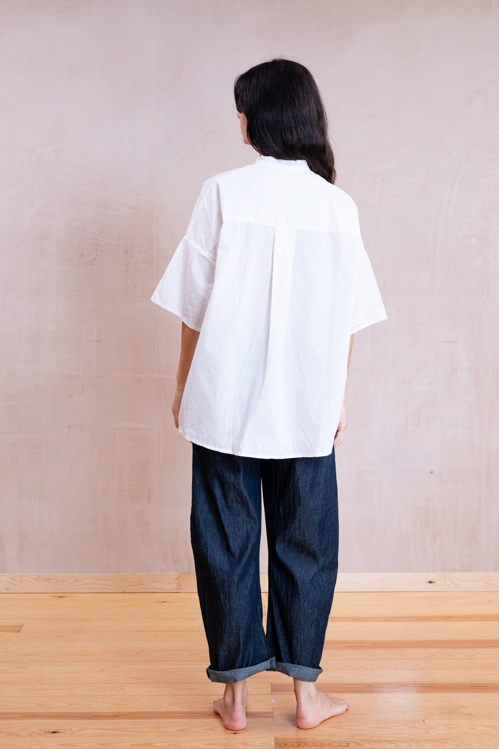 A person shown from the back wearing an oversized white short-sleeve shirt and dark blue denim pants, standing barefoot on a light wood floor against a textured beige wall.