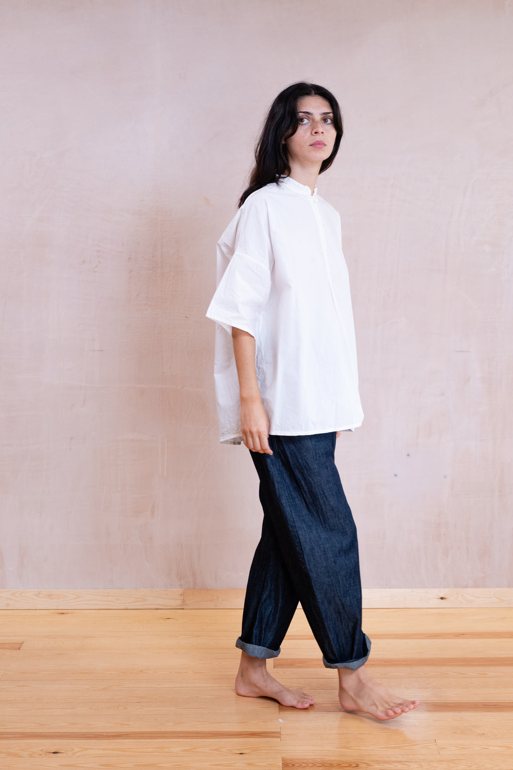 A woman stands barefoot in profile wearing an oversized white short-sleeve top and dark blue cuffed jeans against a light beige wall and wooden floor.