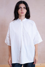 A woman wearing an oversized white short-sleeve button-up shirt with a high folded collar, paired with dark jeans, standing against a textured beige wall.