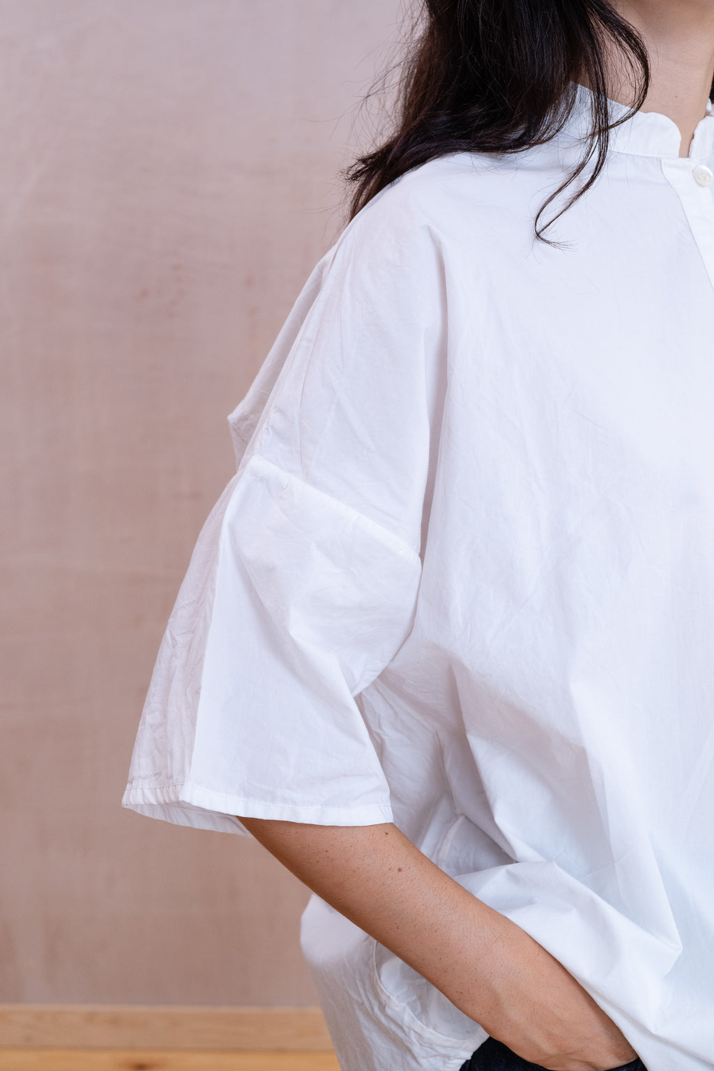 A close-up side view of a person wearing an oversized white short-sleeve blouse or shirt against a soft beige wall.