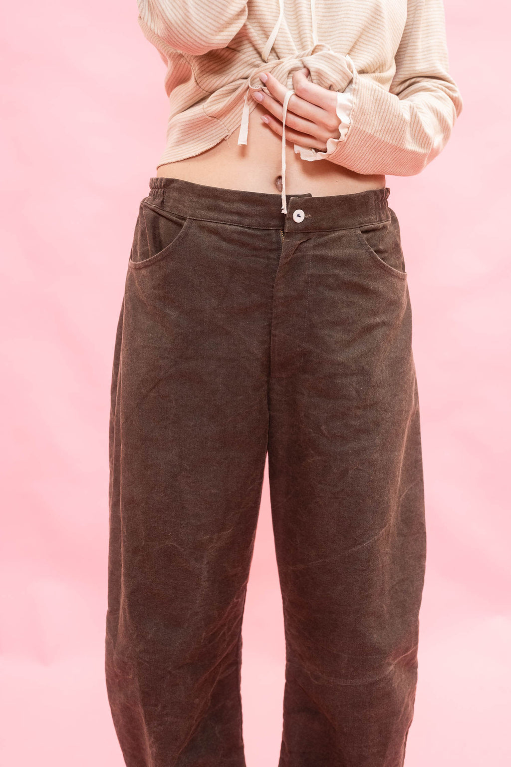 A woman wearing dark brown cotton patina, elasticated waist tapered-leg pants with a wooden button fastening and a cream striped long-sleeve jersey top, standing against a solid pink background.