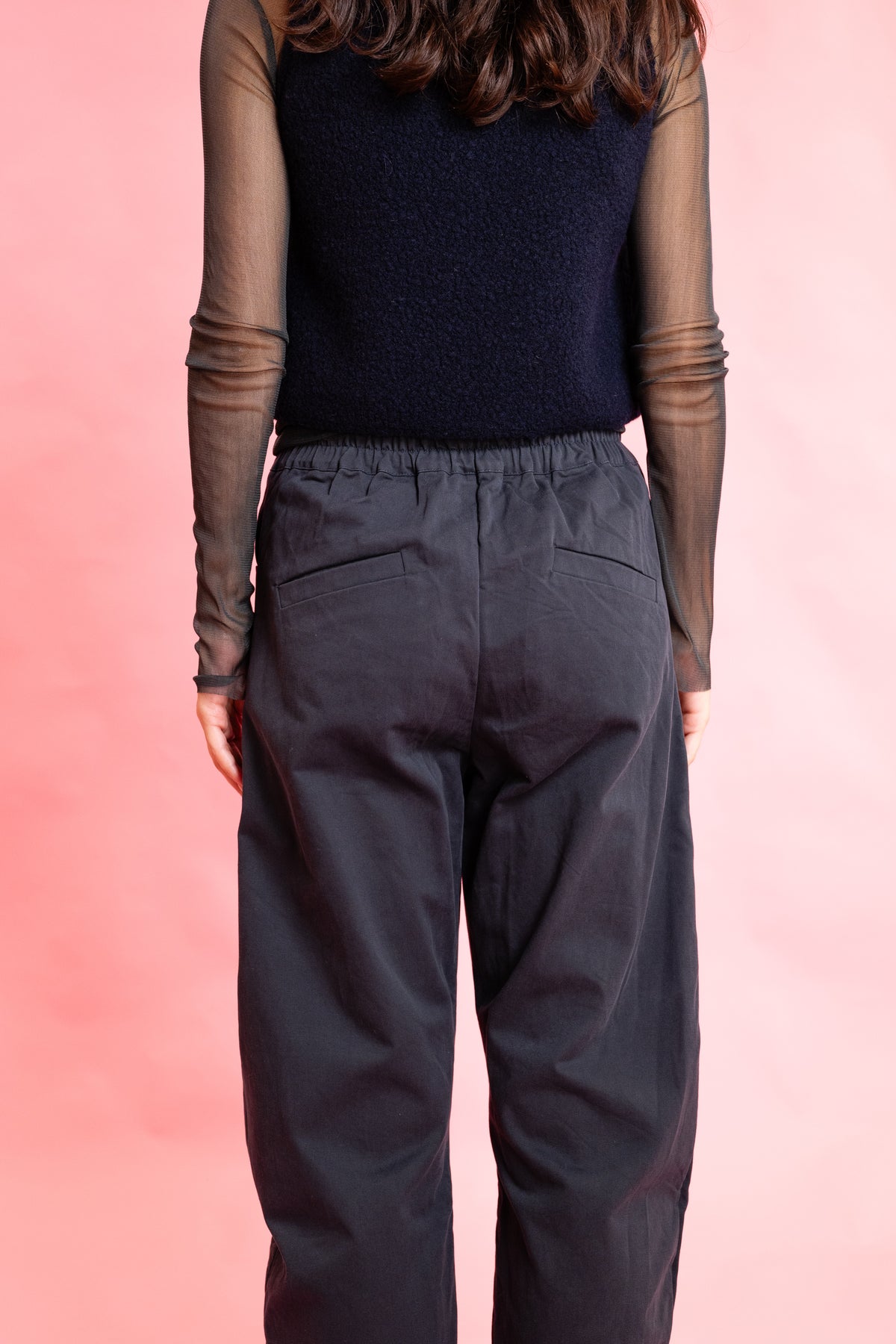 Rear view of a person wearing dark navy wide-leg trousers with an elastic waistband and side pockets, layered over a sheer long-sleeve top and a dark knit sweater, against a solid pink background.
