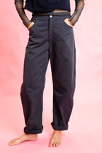 A person wearing loose, dark navy pants with rolled cuffs and side pockets, standing barefoot against a solid pink studio background.