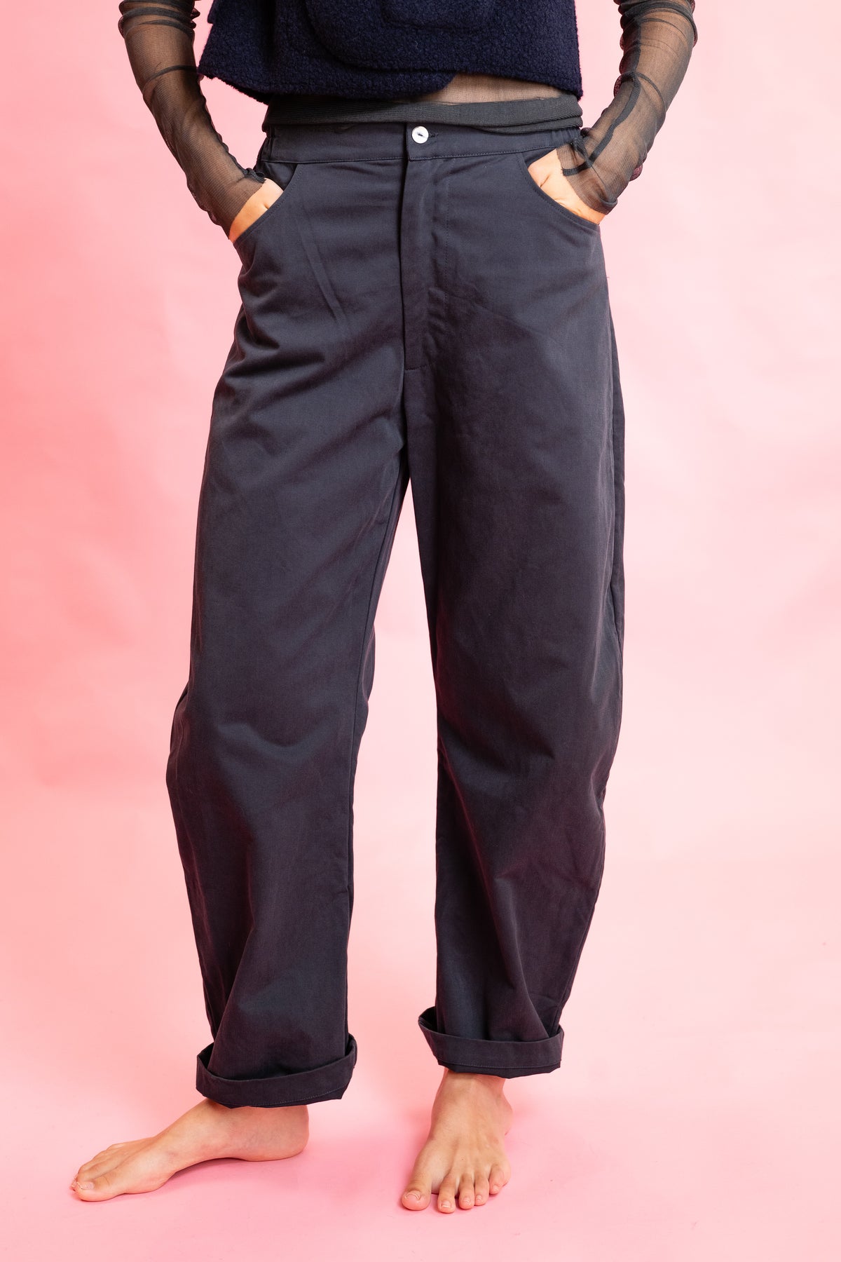 A person wearing loose, dark navy pants with rolled cuffs and side pockets, standing barefoot against a solid pink studio background.
