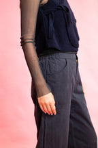 A close-up side view of a person wearing dark navy wide-leg trousers and a dark navy textured top, with sheer black long sleeves, set against a solid pink background.