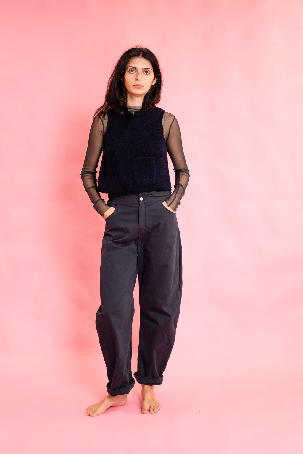 A model stands barefoot against a solid pink studio backdrop, wearing dark navy wide-leg pants, a dark navy velvet vest over sheer black long-sleeve mesh sleeves, and a black top.