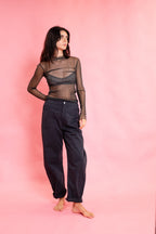 A woman stands barefoot against a solid pink studio backdrop, wearing a sheer black long-sleeve mesh top over a black bralette and loose dark navy pants.