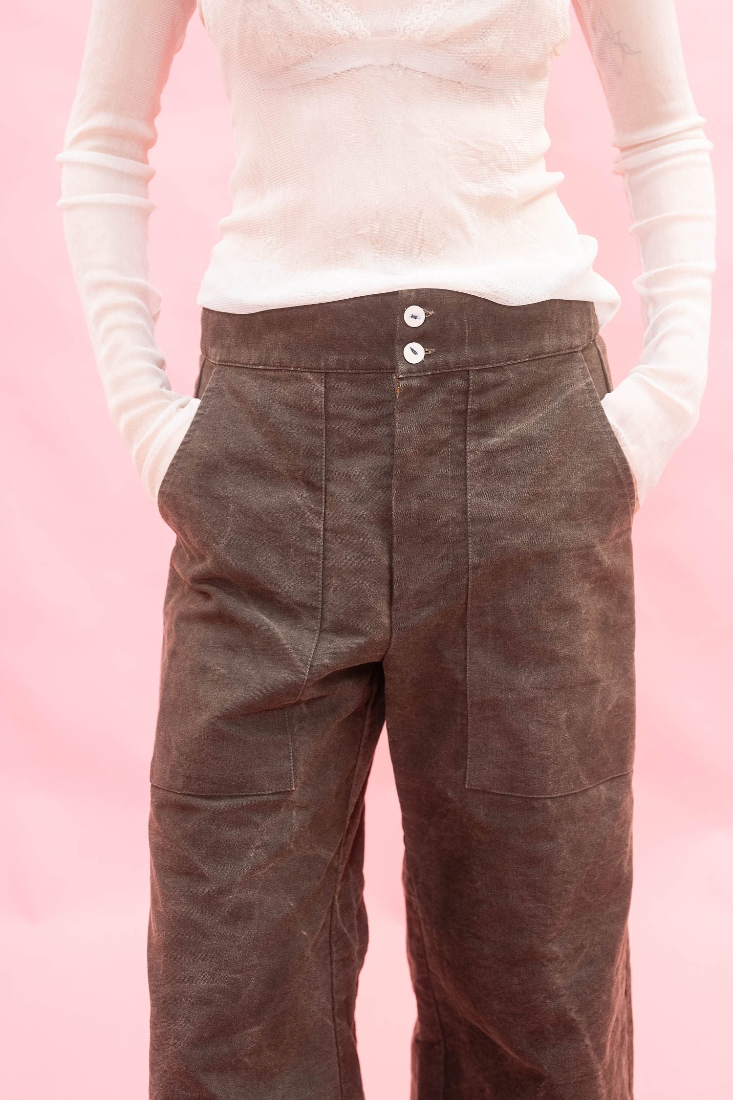 A person wearing dark brown wide-leg cotton pants with a white long-sleeve top, standing against a solid pink background.