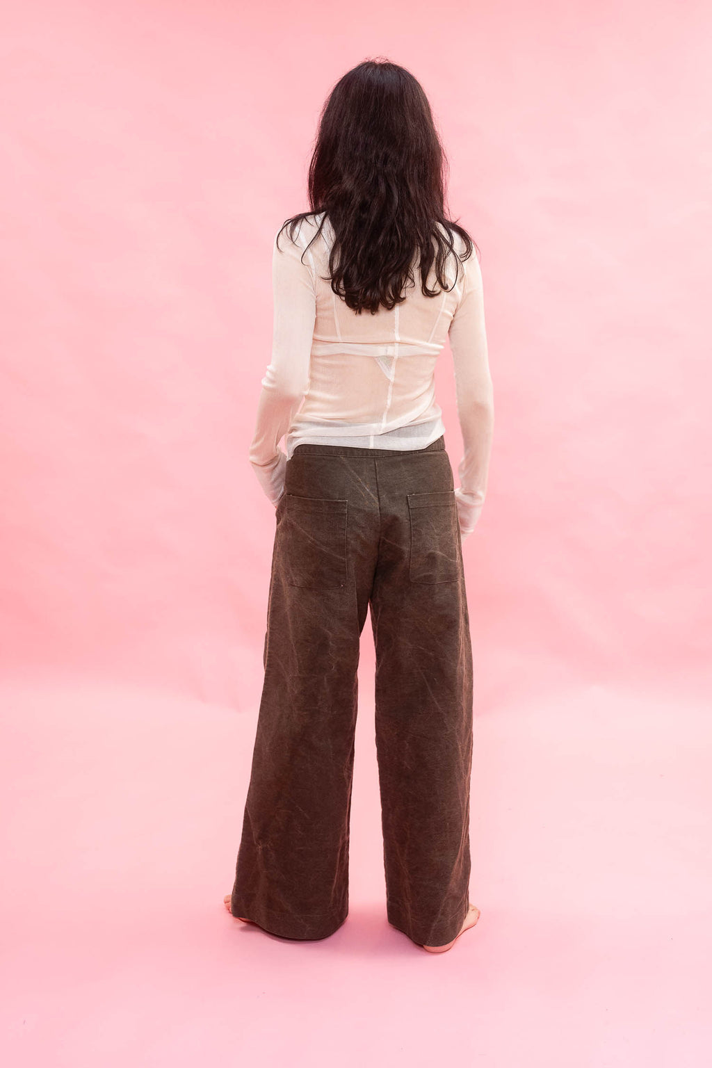 A woman viewed from behind wearing a white long-sleeve top and dark brown wide-leg cotton pants, standing against a solid pink studio backdrop.