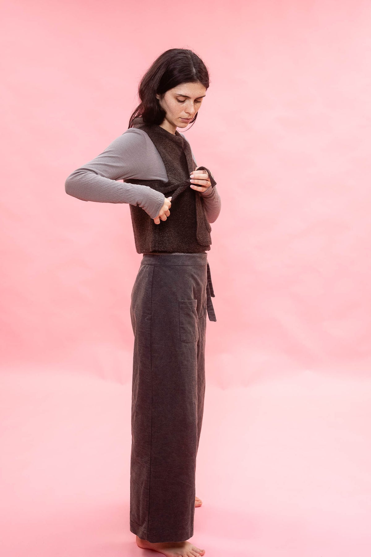 A woman modeling a dark brown wool vest over a gray long-sleeve shirt and dark navy wide-leg linen pants, standing against a solid pink studio background.