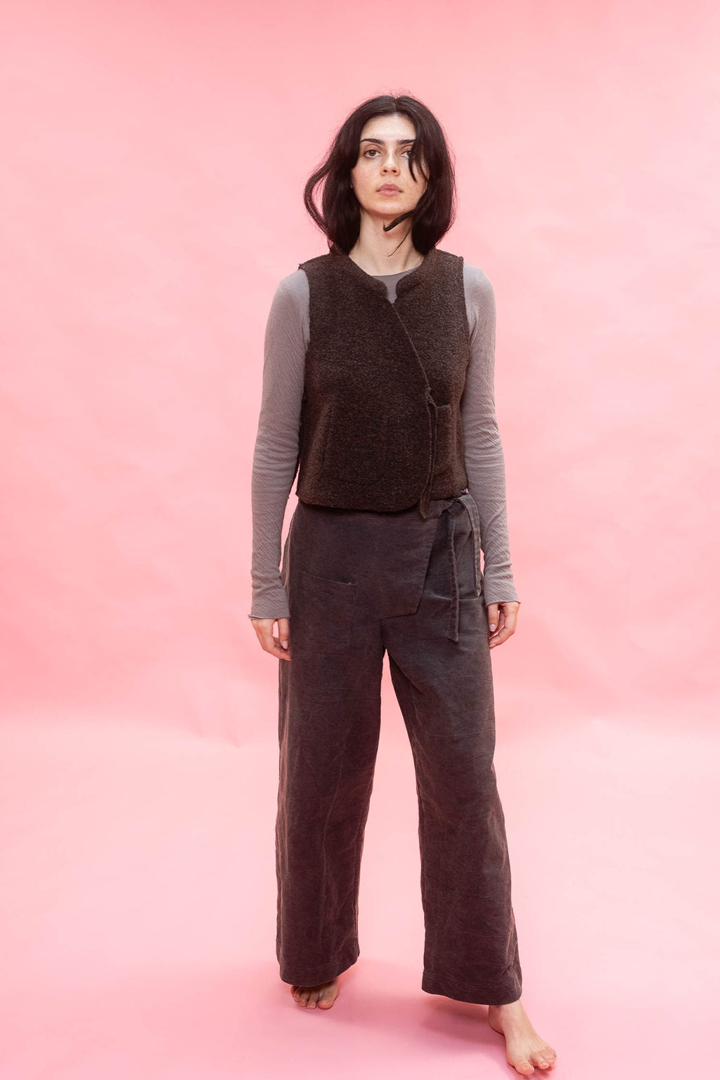A woman stands barefoot against a solid pink studio backdrop, wearing a dark brown sleeveless textured vest over a gray long-sleeve top and dark navy wide-leg pants.