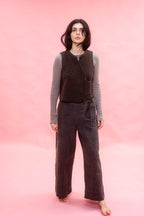 A woman stands barefoot against a solid pink studio backdrop, wearing a dark brown sleeveless textured vest over a gray long-sleeve top and dark navy wide-leg pants.