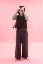 A person stands barefoot against a solid pink backdrop, wearing a dark brown sleeveless textured vest over a light gray long-sleeve top and dark navy wide-leg pants.
