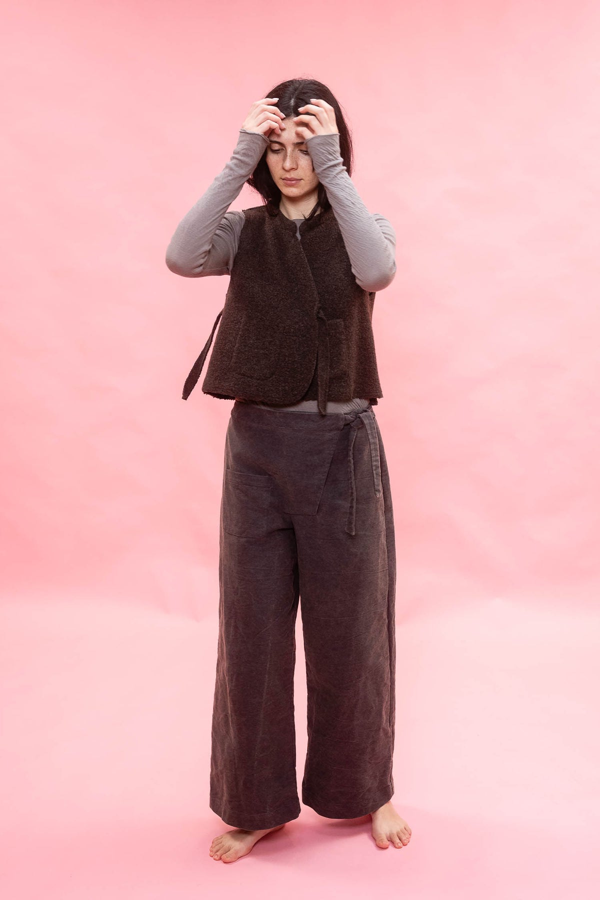 A person stands barefoot against a solid pink backdrop, wearing a dark brown sleeveless textured vest over a light gray long-sleeve top and dark navy wide-leg pants.