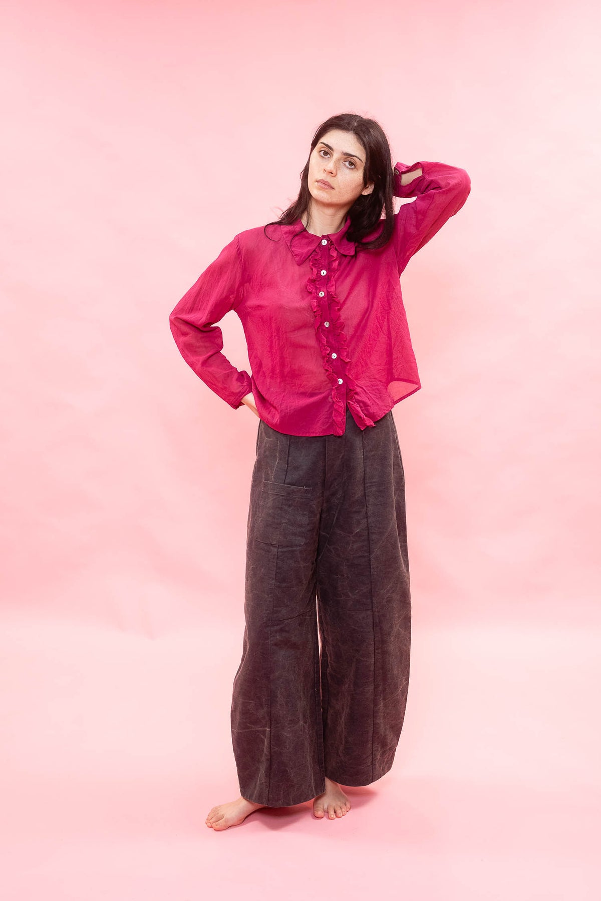 Person wearing a pink sheer blouse and brown pants on a pink background