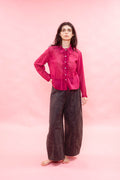 A woman stands barefoot against a solid pink studio backdrop, wearing a bright pink long-sleeve button-front shirt and dark charcoal wide-leg pants.