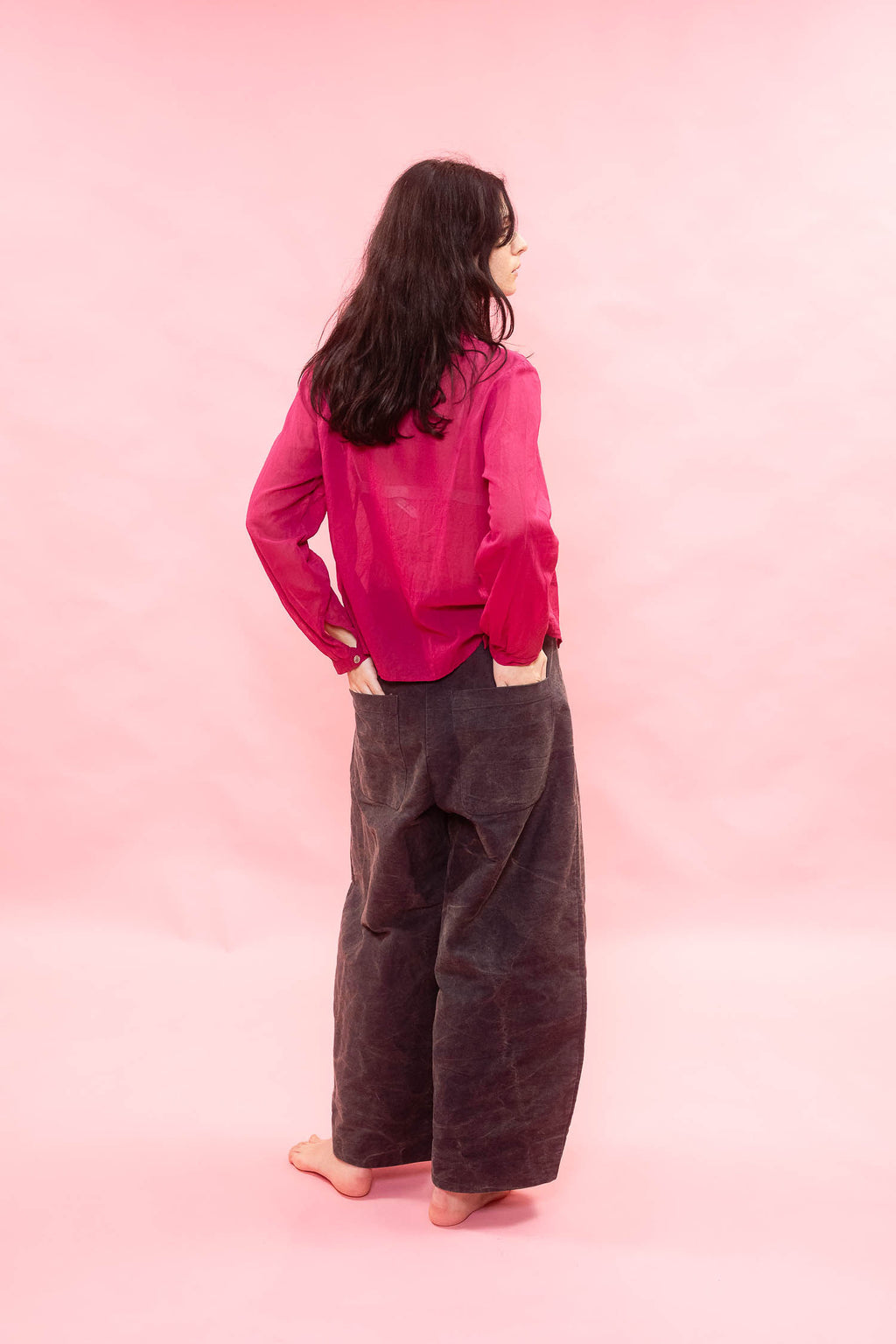 A woman stands barefoot against a solid pink backdrop, viewed from behind. She is wearing a bright pink long-sleeve top and dark brown wide-leg pants.