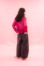 A woman stands barefoot against a solid pink backdrop, viewed from behind. She is wearing a bright pink long-sleeve top and dark brown wide-leg pants.