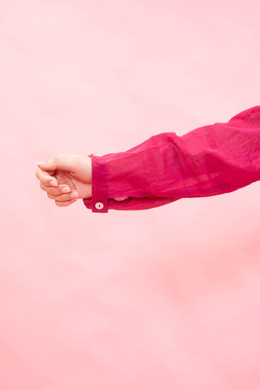 A close-up of a person's hand and forearm wearing a bright pink long-sleeve button-up shirt against a solid light pink background.