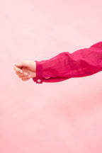 A close-up of a person's hand and forearm wearing a bright pink long-sleeve button-up shirt against a solid light pink background.