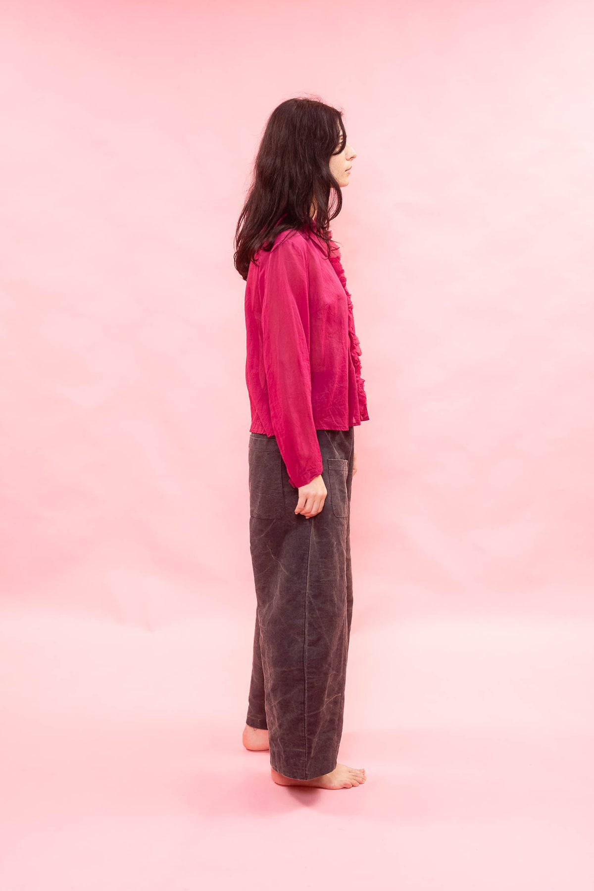 A woman stands in profile against a solid pink studio backdrop, wearing a bright pink long-sleeve button-front top and dark brown wide-leg pants.