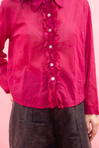 A close-up of a person wearing a bright pink long-sleeve blouse with a ruffled button placket and dark brown pants, set against a solid pink background.