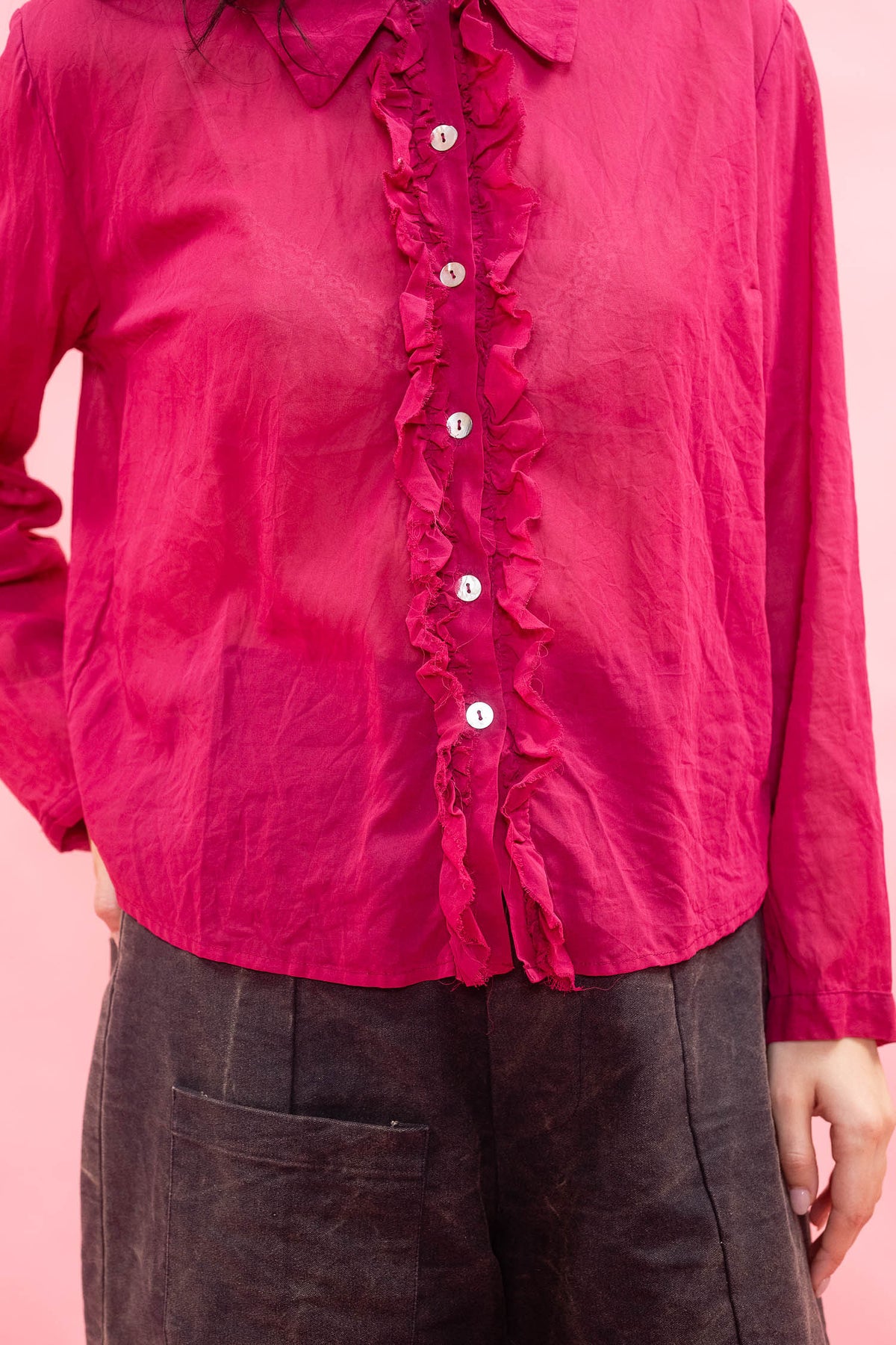 A close-up of a person wearing a bright pink long-sleeve blouse with a ruffled button placket and dark brown pants, set against a solid pink background.