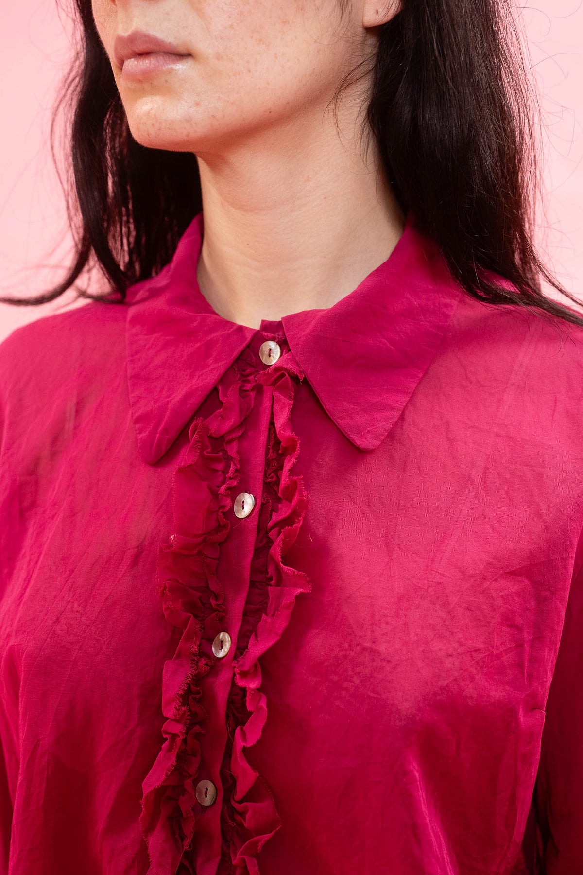 A close-up of a model wearing a vibrant magenta pink blouse with a ruffled button-down front and a classic collar, set against a solid pink background.