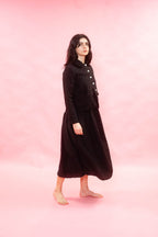 A woman stands barefoot against a solid pink studio background, wearing a black long-sleeve button-front jacket with a high collar and a matching black midi skirt.